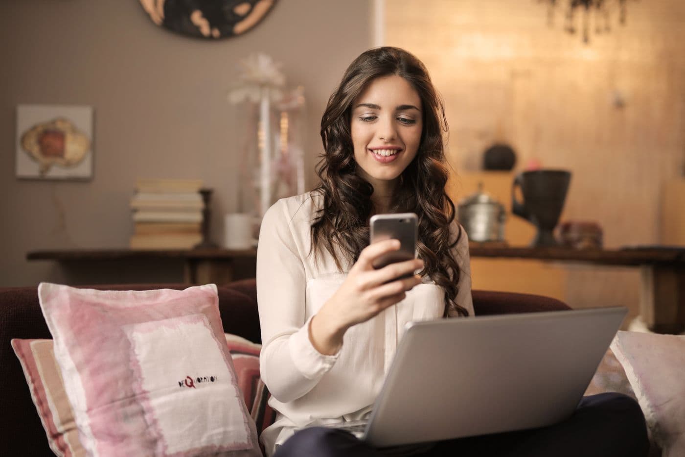 Wanita tersenyum sambil membalas pesan di ponsel dengan laptop terbuka di pangkuannya