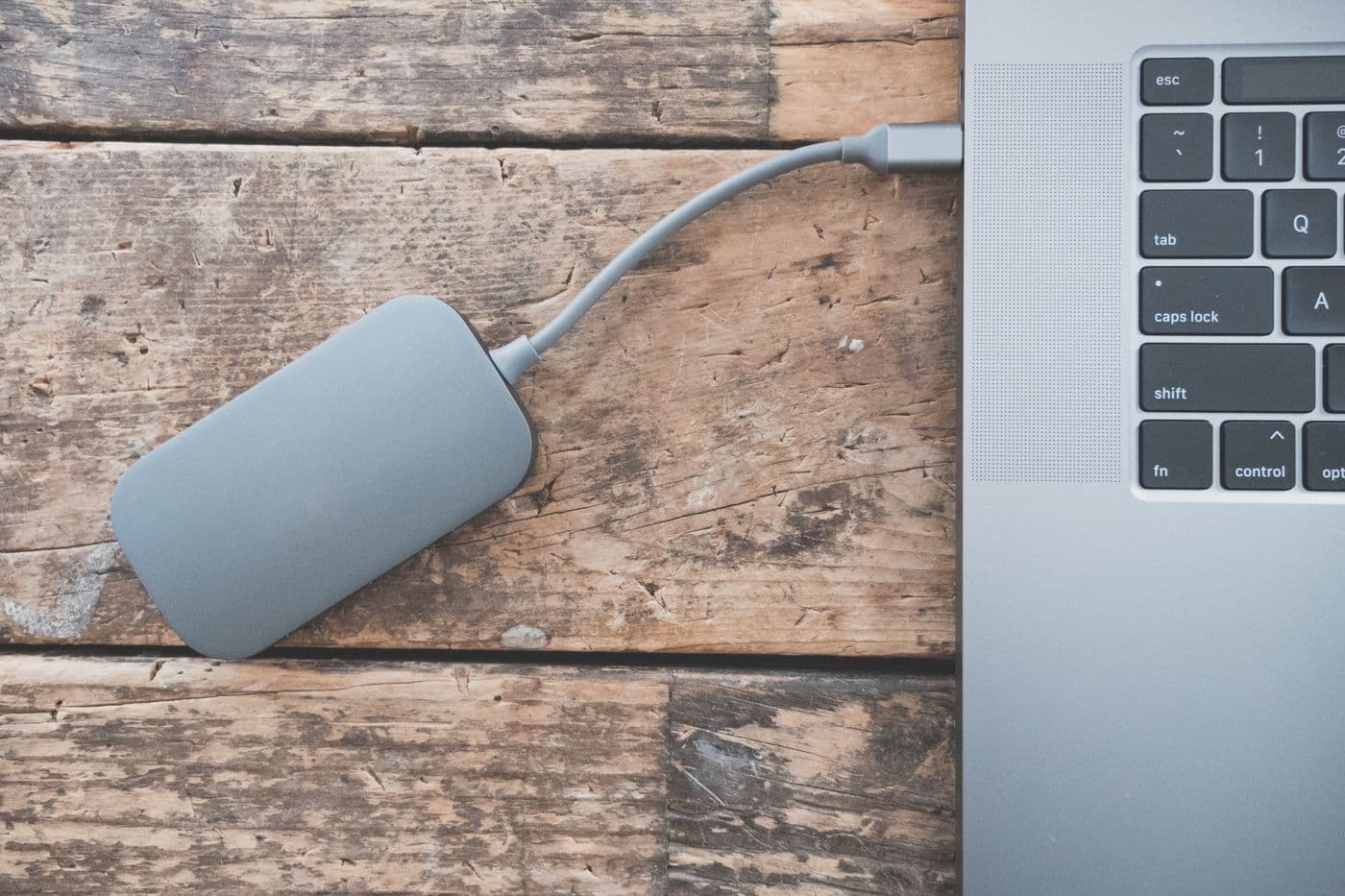 Hard disk eksternal abu-abu tersambung kabel ke laptop di atas meja kayu