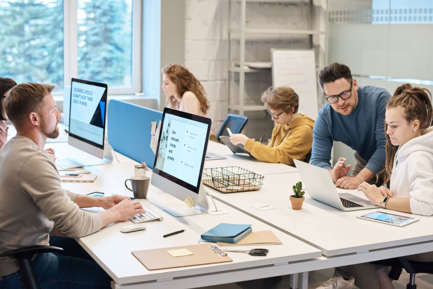 Pria berkacamata membimbing rekan kerja perempuan di depan laptop di ruang kantor yang ramai