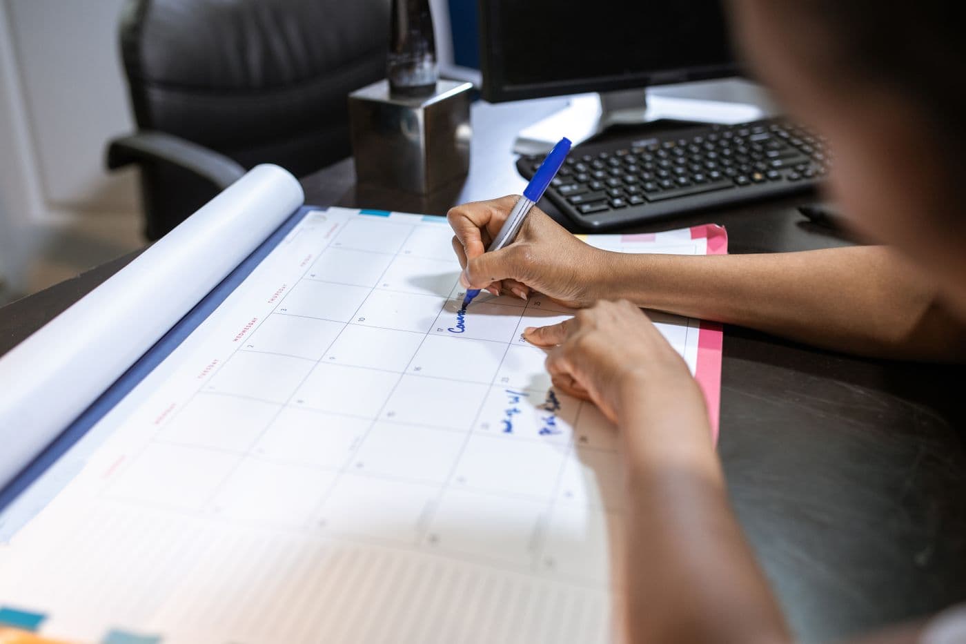 Tangan menulis jadwal dengan spidol biru di kalender meja besar, dengan keyboard dan monitor di latar belakang