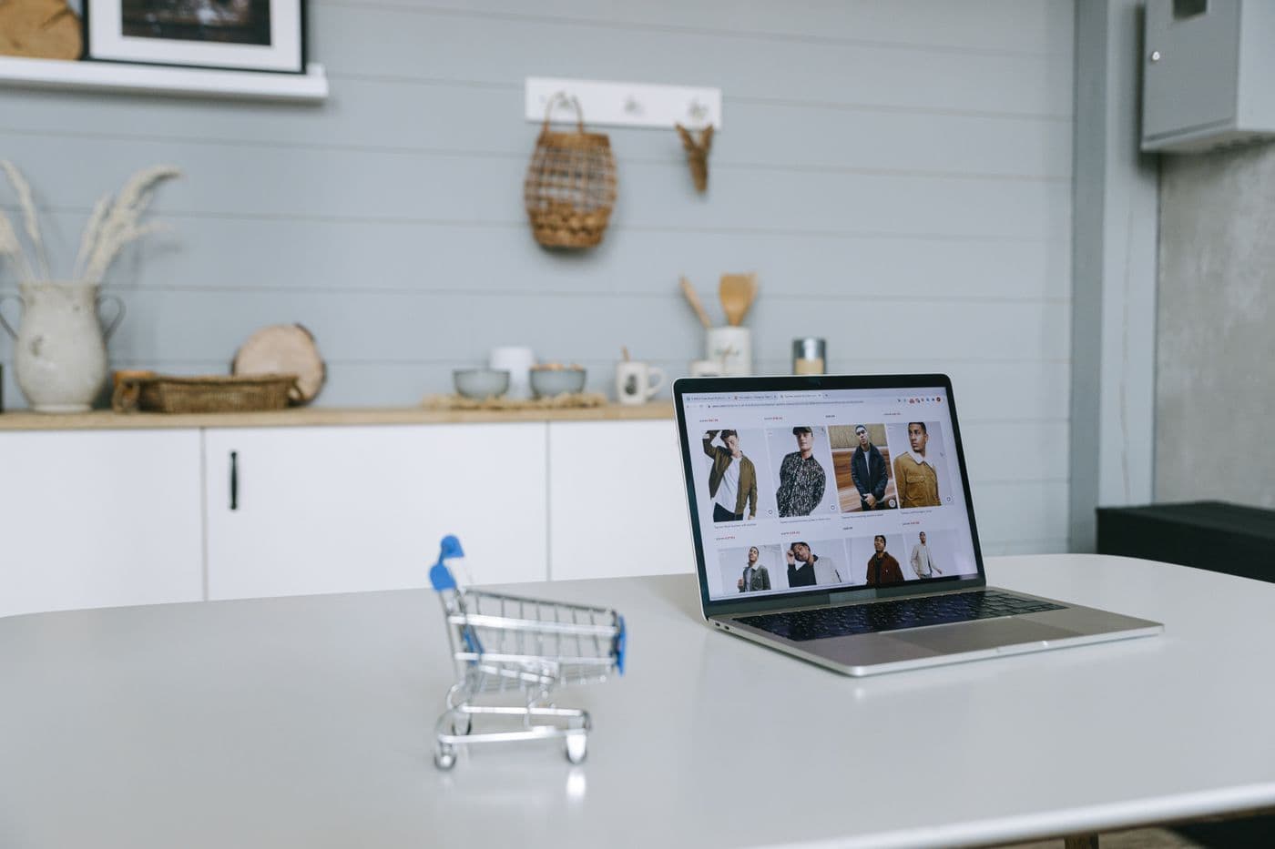Troli belanja mini kosong di meja putih, di belakangnya laptop terbuka menampilkan katalog toko online