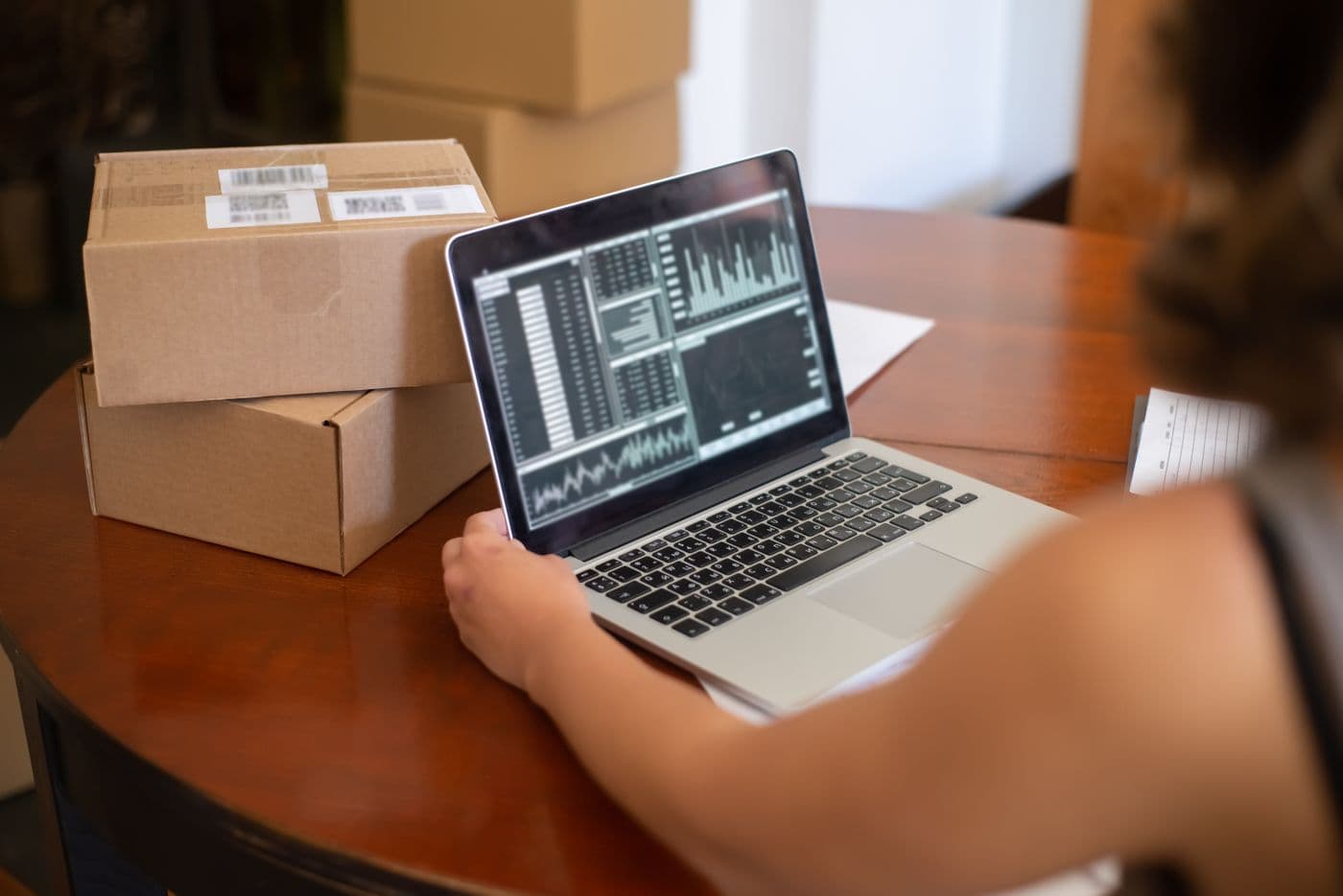 Seseorang memeriksa grafik data penjualan di layar laptop dengan tumpukan kotak paket di sampingnya