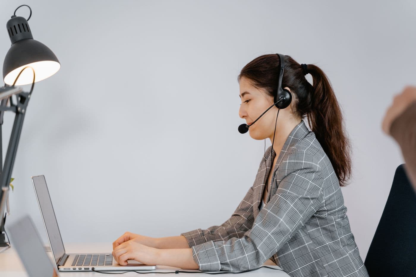Perempuan memakai headset dengan mikrofon sedang mengetik di laptop di meja kerja