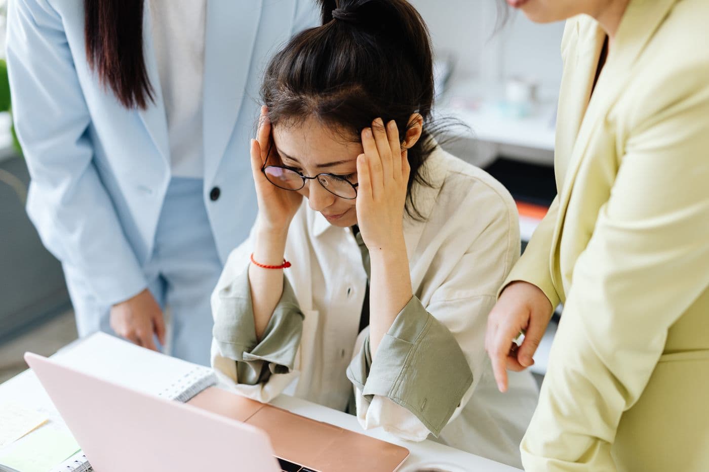 Perempuan berkacamata memegang kepala dengan cemas di depan laptop, dua rekan berdiri di sampingnya menunjuk layar