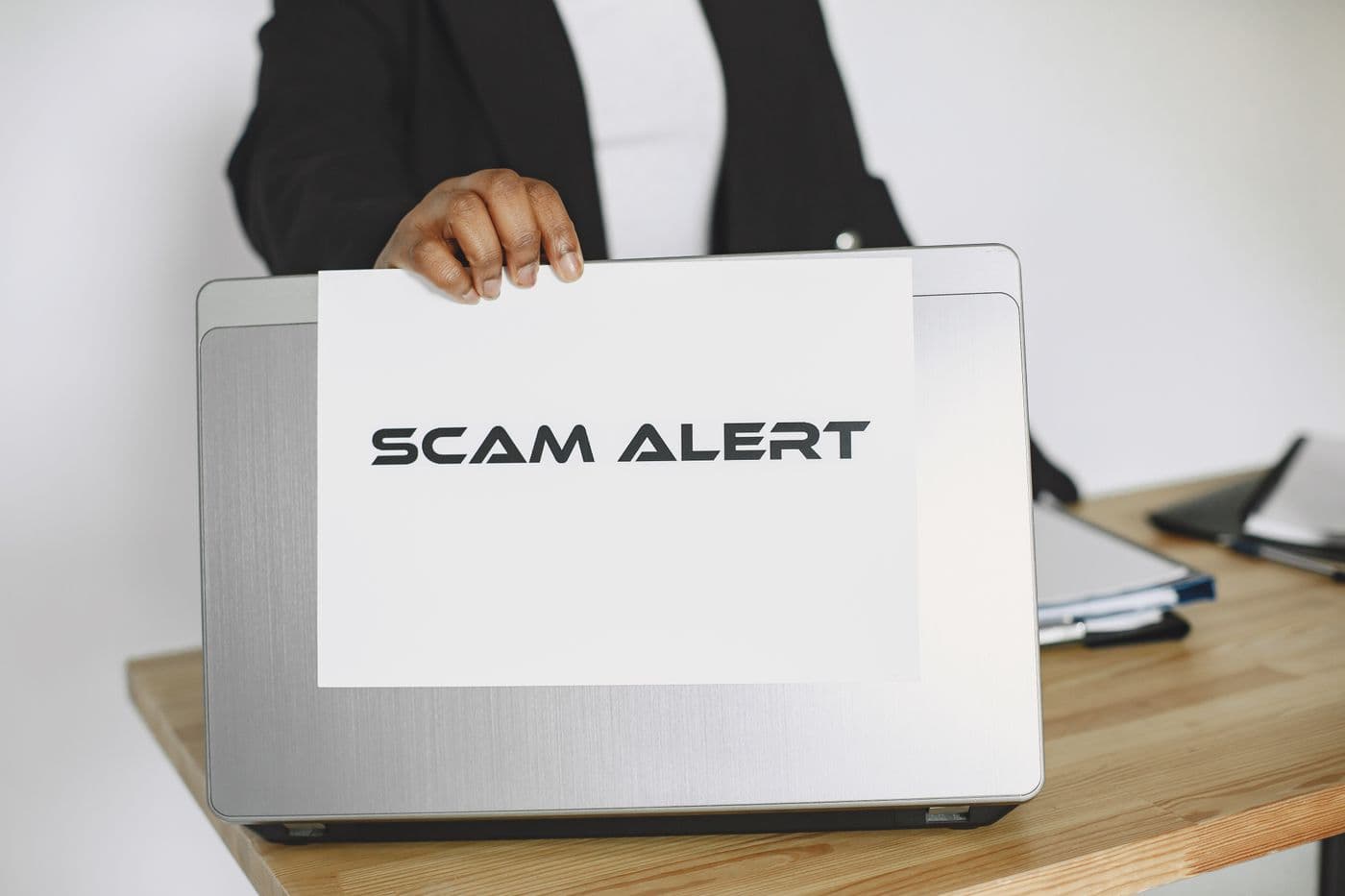 Tangan memegang kertas bertuliskan Scam Alert di depan laptop yang tertutup di atas meja kayu